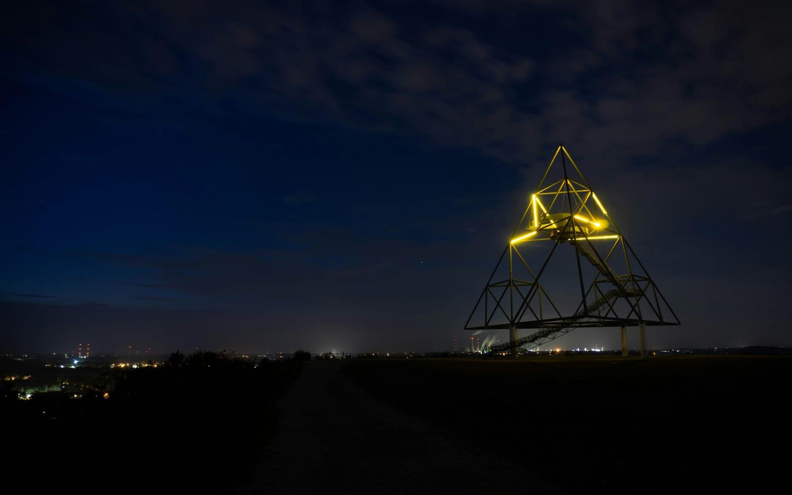 Der beleuchtete Tetraeder auf der Halde Beckstraße in Bottrop bei Nacht, im Hintergrund die Lichter des Ruhrgebiets